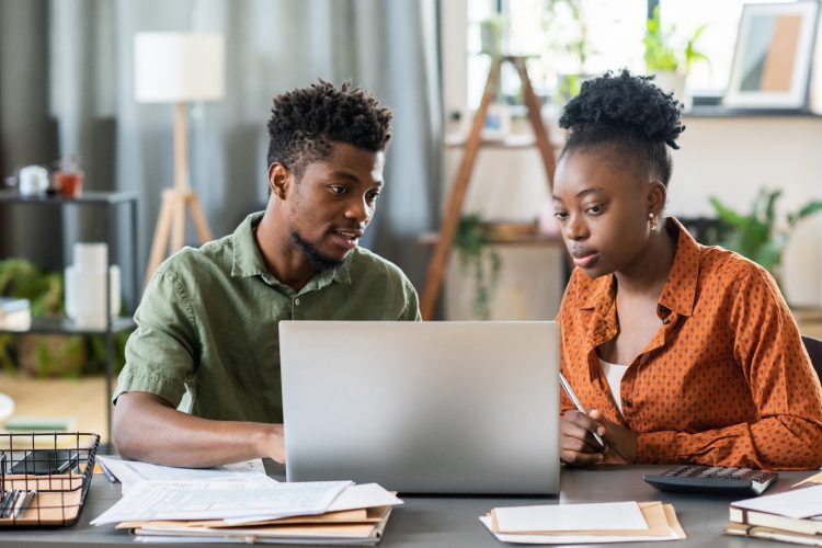 Young blackman sitting at table with bills and document and examining online tax form with girlfriend on laptop